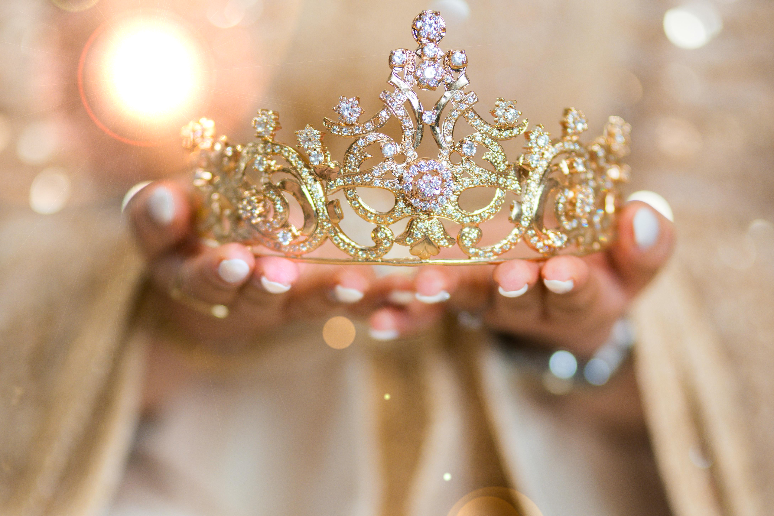 Im Vordergrund sieht man eine goldfarbene Tiara, gehalten von zwei Händen. Die Person, von der man nur die Hände sieht, trägt ein beige-farbenes Gewand.