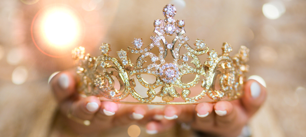 Im Vordergrund sieht man eine goldfarbene Tiara, gehalten von zwei Händen. Die Person, von der man nur die Hände sieht, trägt ein beige-farbenes Gewand.