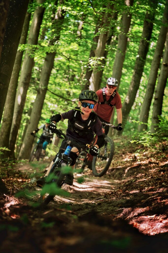 Im Vordergrund sieht man ein Kind auf einem Mountainbike, im Hintergrund einen Erwachsenen auf einem Mountainbike. Sie fahren den Trail im Wald entlang. Ganz hinten, verschwommen, ist noch eine Person auf einem Mountainbike zu sehen.